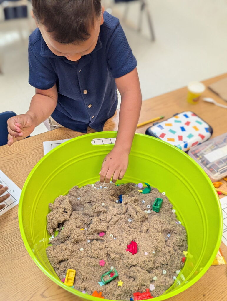 A child in a blue shirt looks for letters in a green tub filled with sand, on a table.