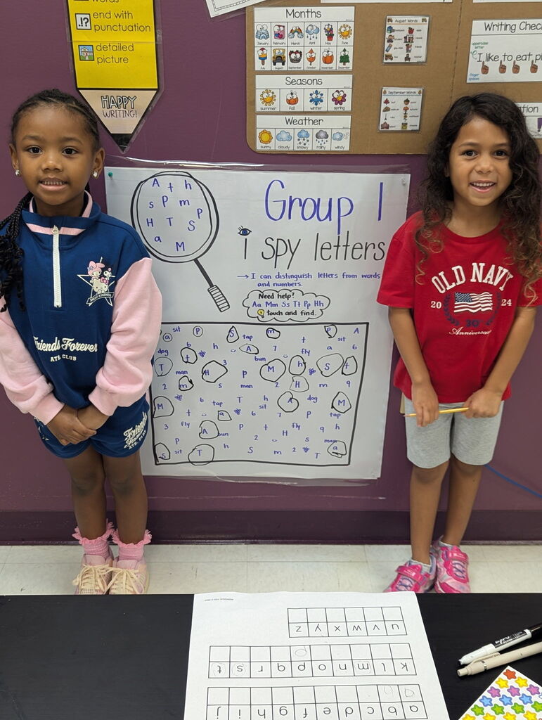 Two young girls stand beside a table displaying a worksheet and a classroom bulletin board with letters and activities.