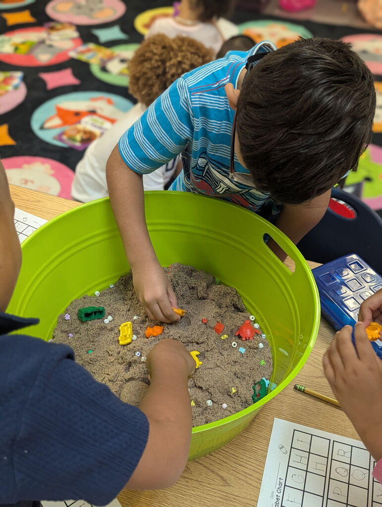 Three children engage in an activity with letters in a green bucket. One child in a striped shirt bends over the bucket.