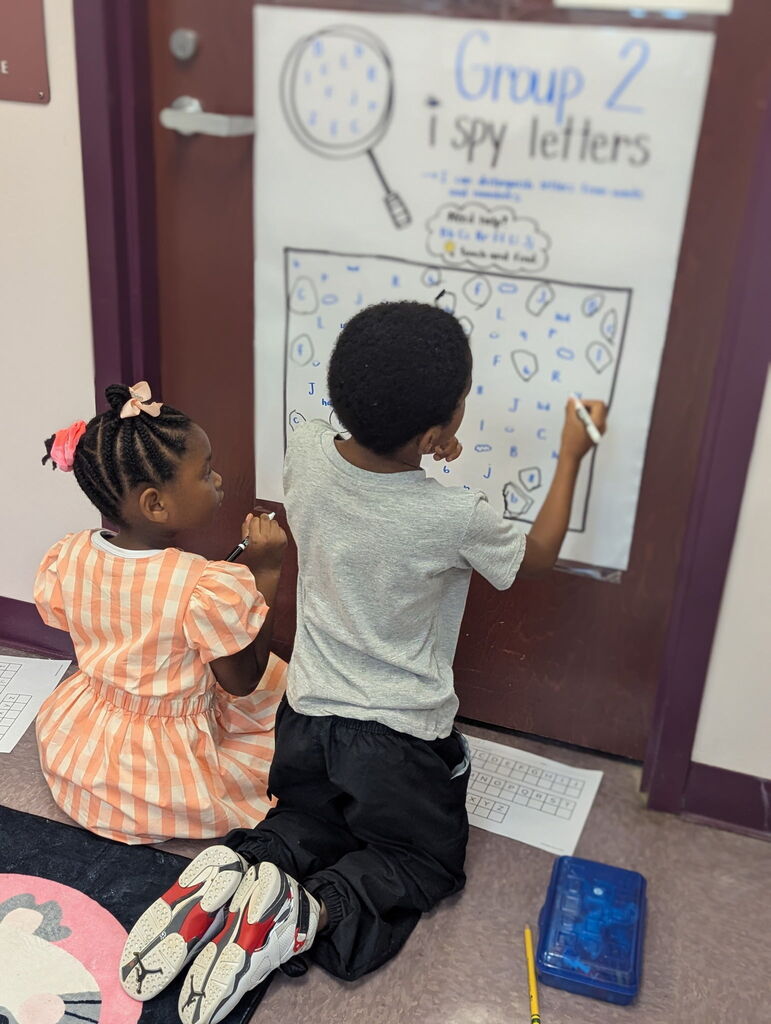 Two young children sit on a floor, writing on a large chart titled "Group 2 i spy letters."