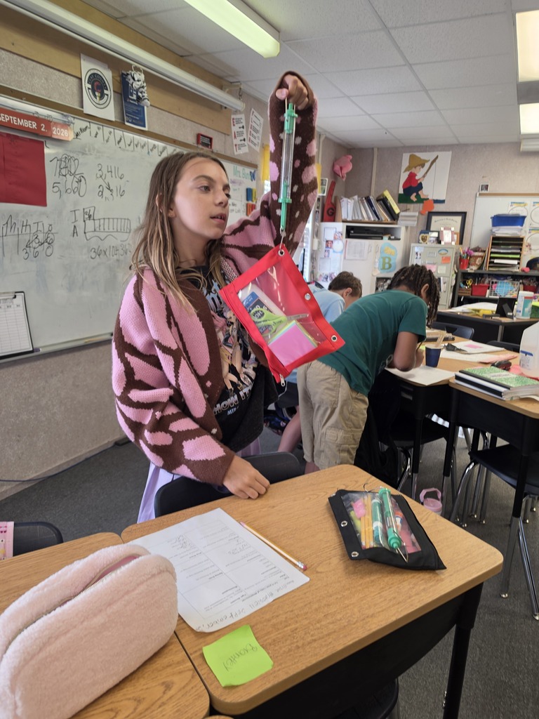 Woman with pink jacket holds up a folder in classroom. Whiteboard behind her. Other students work at desks in background.