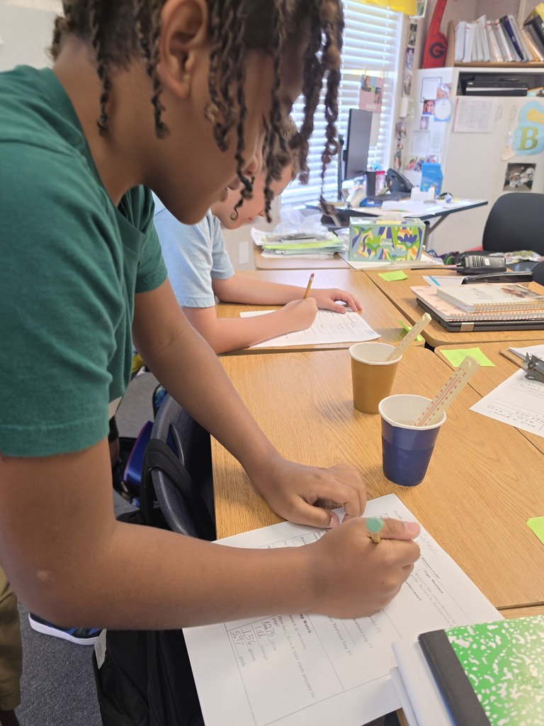 A person wearing a green shirt, seated at a desk, writing on paper. Behind them is a blurred figure also writing.