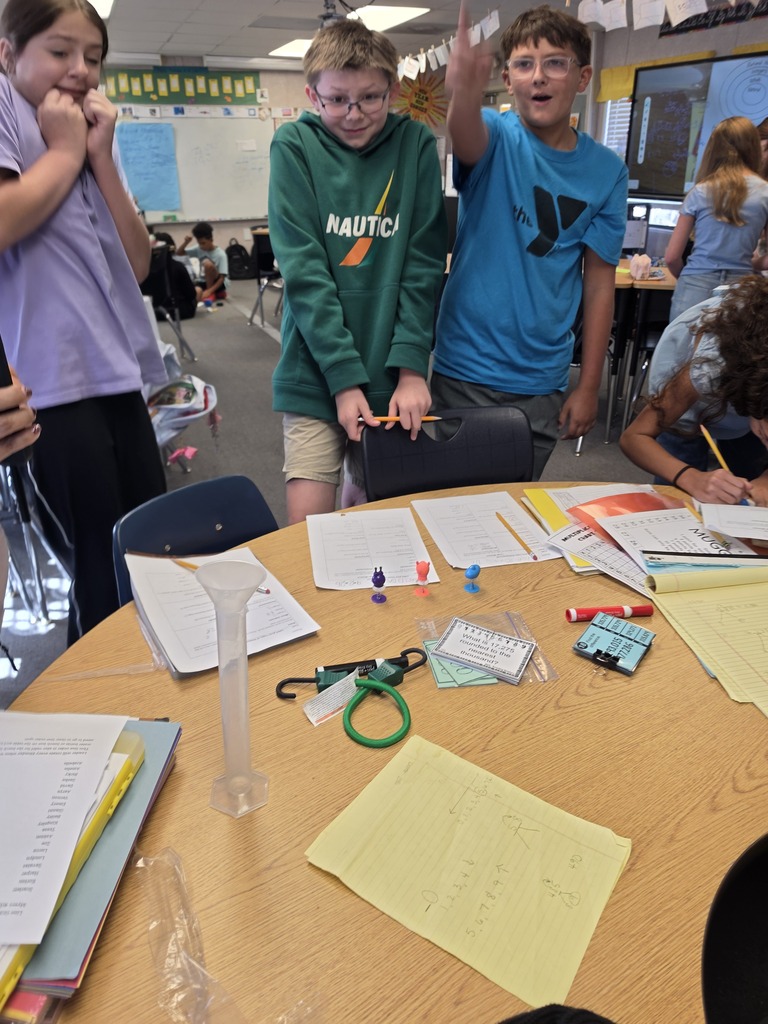 Four children standing at a table in a classroom. The table holds books, scissors, and papers.