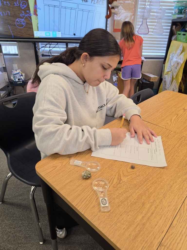 A young person, wearing a white hoodie, sits at a table with science equipment, writing on paper.