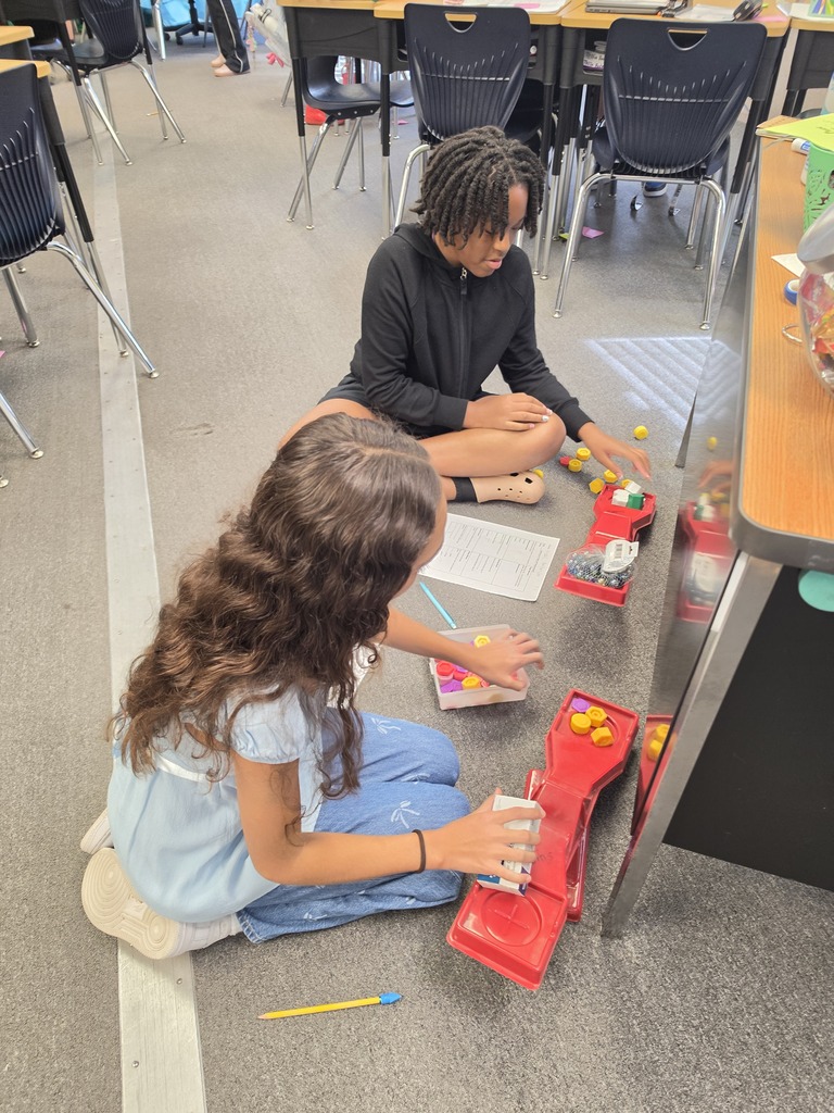 Two children sit on the floor in a classroom, working on a project with colorful toys.
