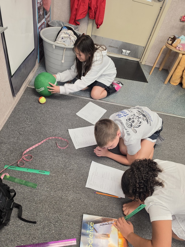 Three students on floor, one holding a ball and two writing. Papers, pencils, and colorful objects scattered.