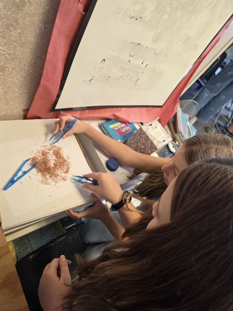 Two people are painting with blue paint and brown powder on a white canvas. A large white paper is above them.