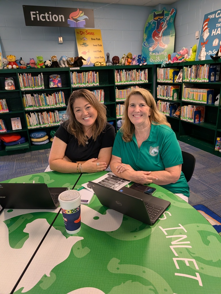 Two women sit at a round table in a classroom. A laptop, a cup, and other items are on the table. Bookshelves are in the background.