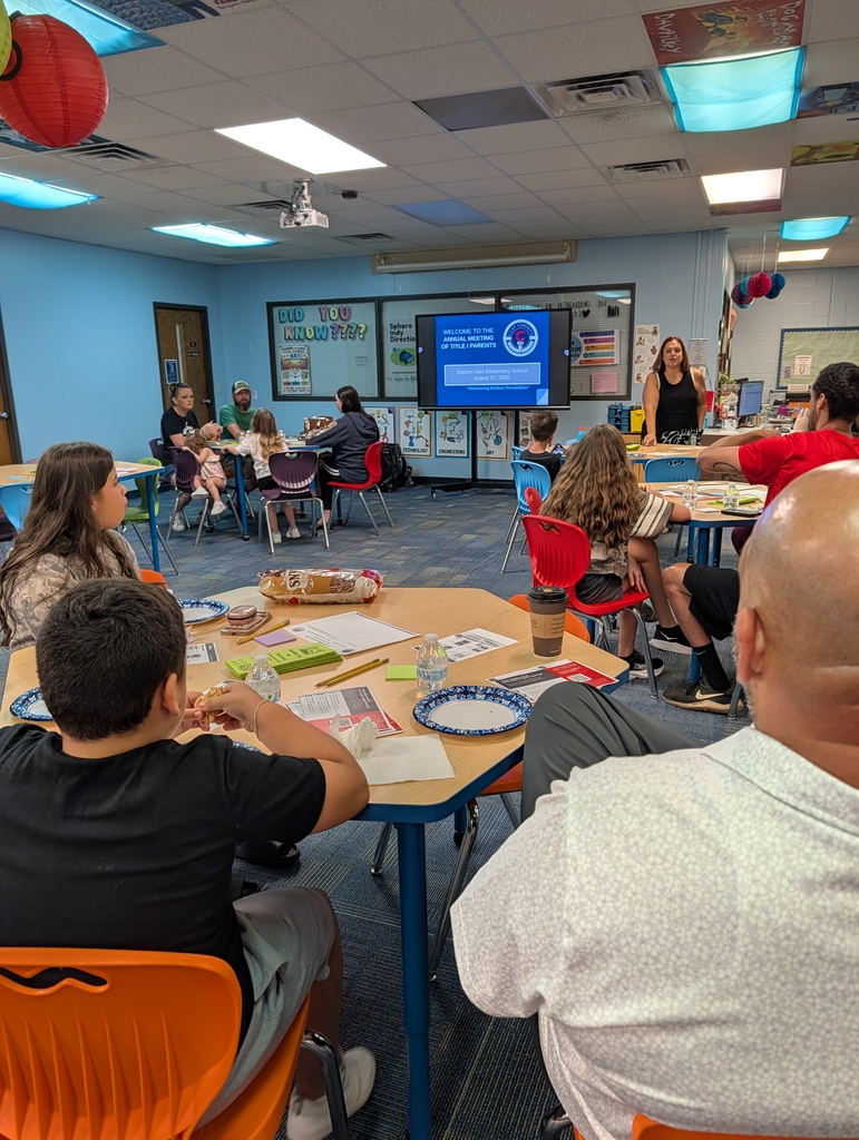 A room with tables and chairs, a projector screen, and adults and children. Some are seated while others stand.