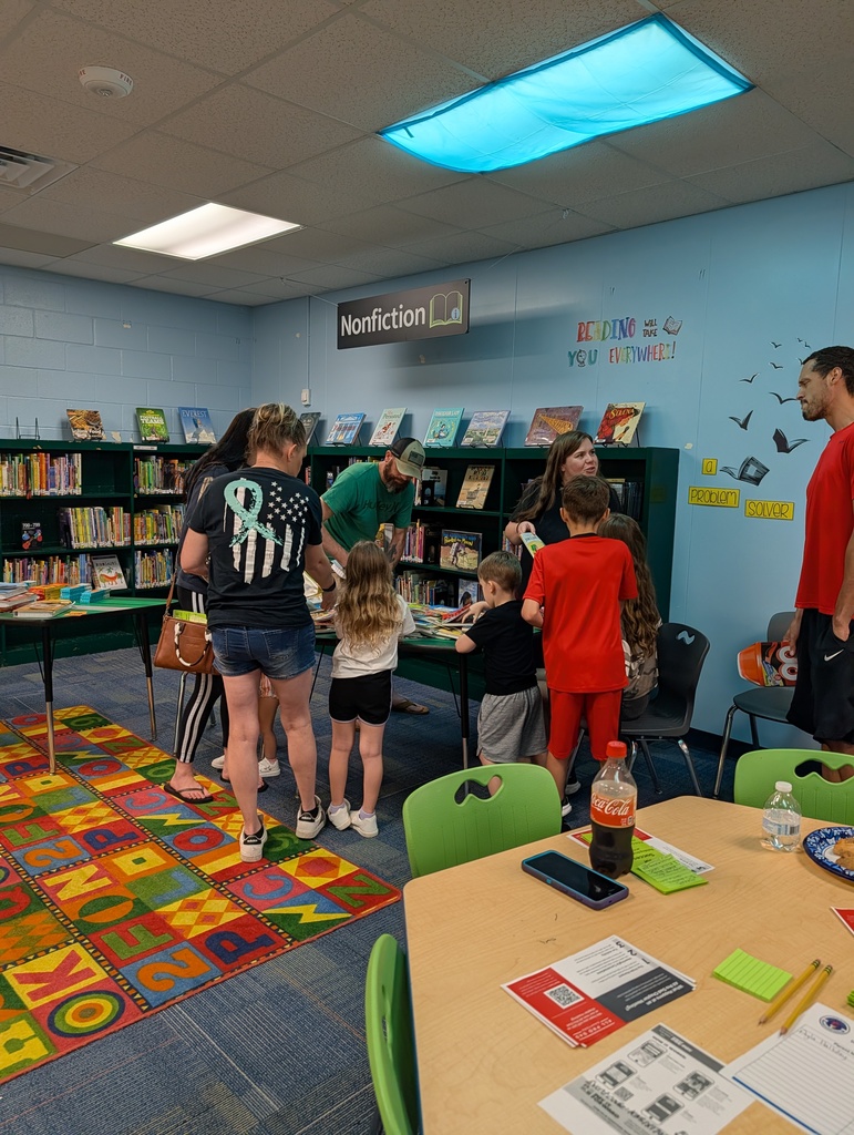 A library with children and adults near shelves. Some are looking at books. A table has a phone, soda, and papers.