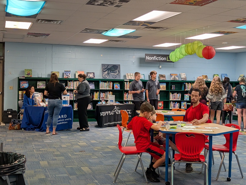 A busy library area with people seated around tables. Shelves filled with books are in the background.