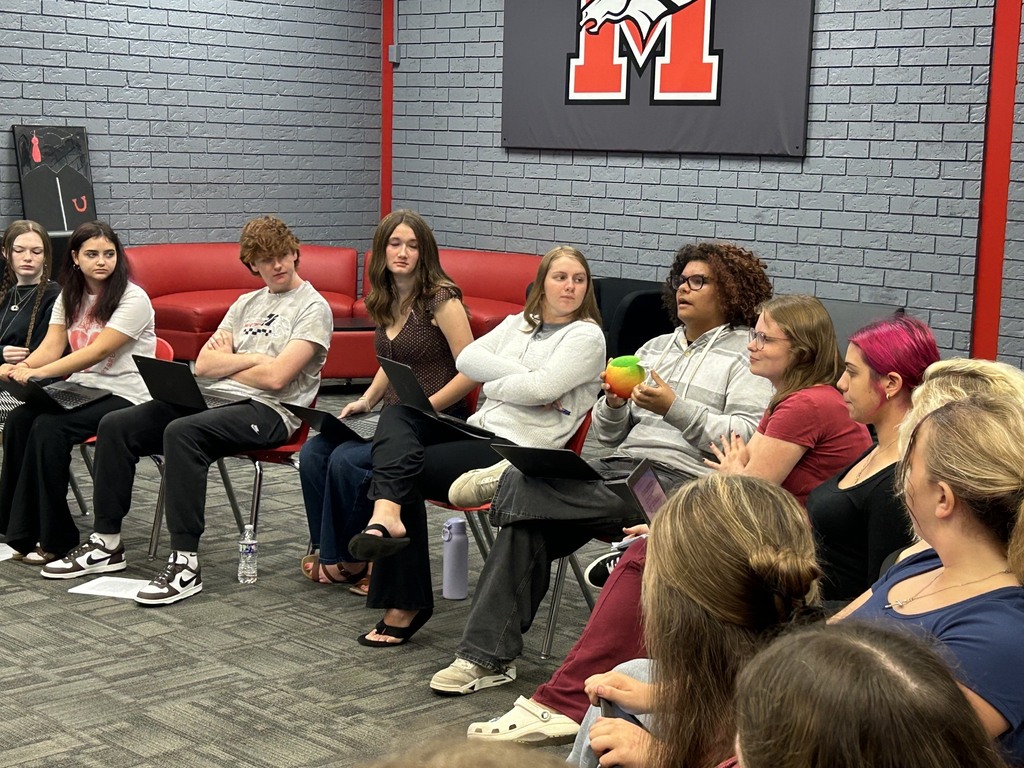 A group of people sit on chairs in a classroom. They have laptops on their laps.
