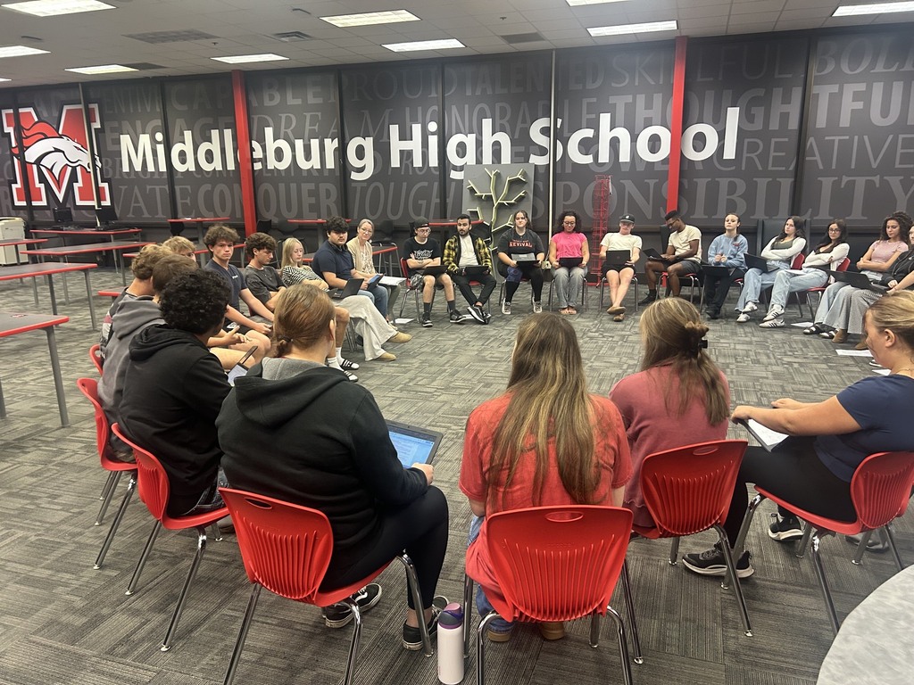 A circle of students and staff seated in red chairs at Middleburg High School. A large wall behind them reads "Middleburg High School."