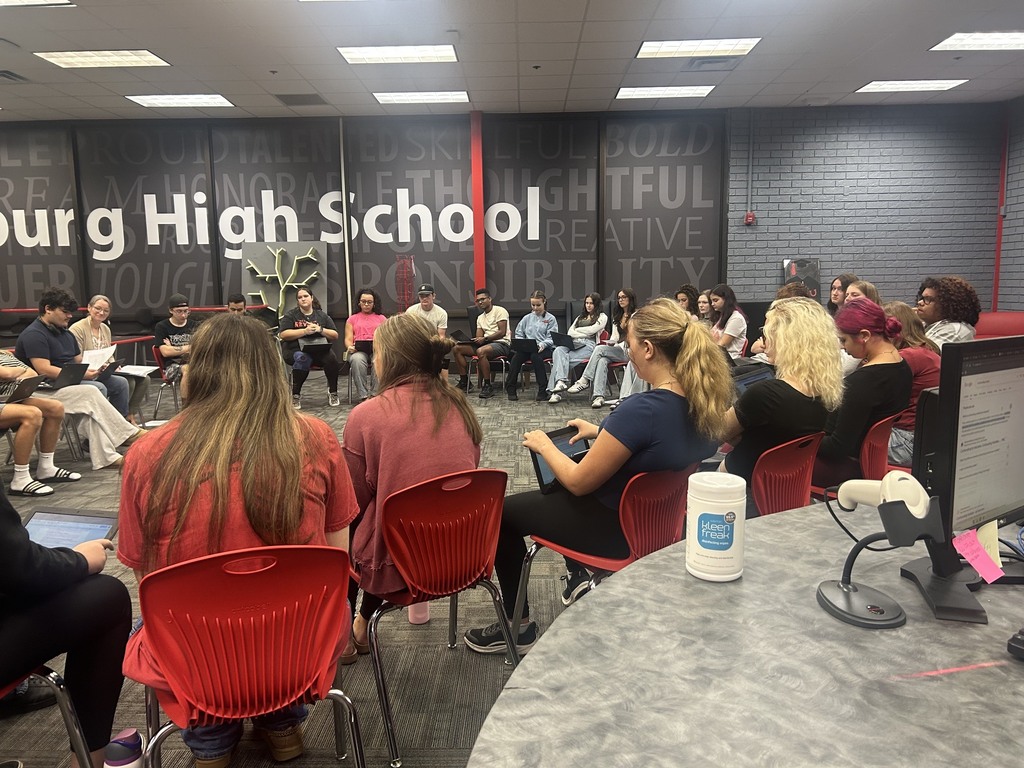 Indoor classroom with students seated in red chairs. A wall reads "High School" and a desk has a computer monitor.