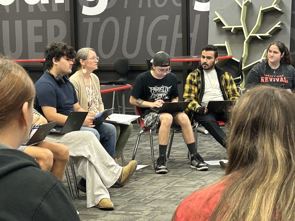 A group of people with laptops are seated in chairs around a room, facing a black wall with bold text.