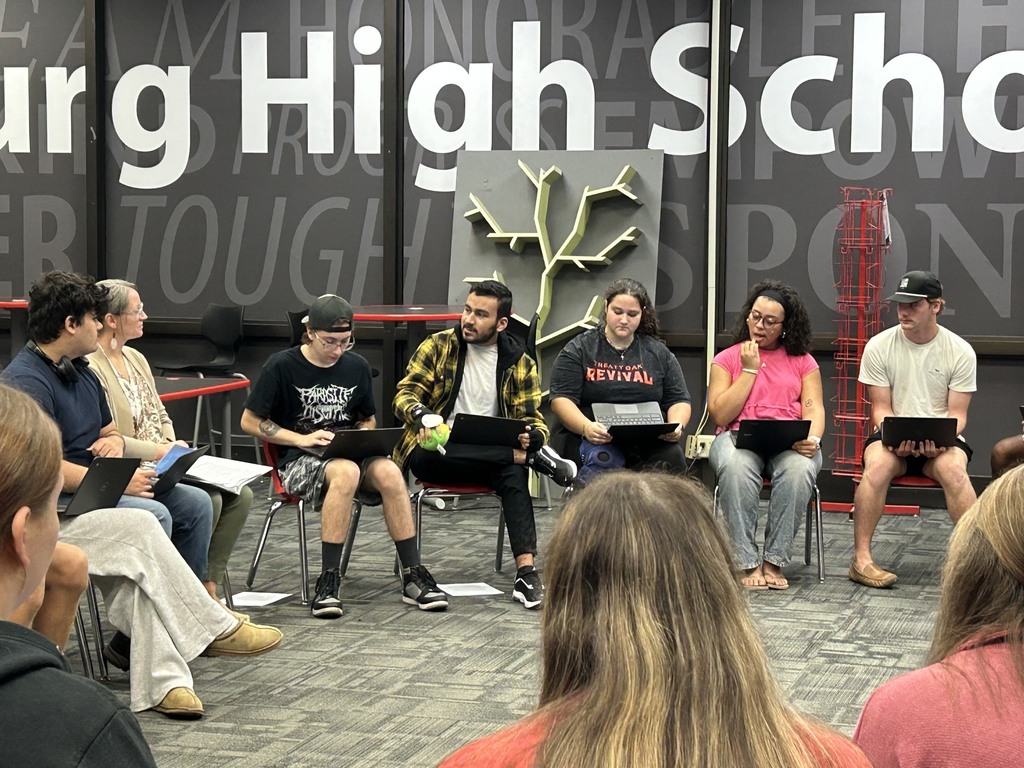 A group of students with laptops sit in chairs around a room, with "Burg High School" on a wall.