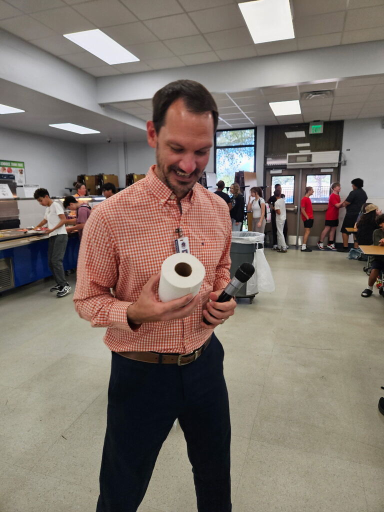 A man holding a microphone and paper roll smiles in a room. Behind him, students walk.