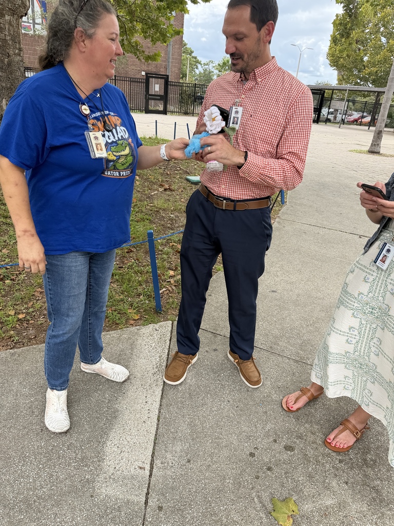 Three people stand outside on a concrete sidewalk. A man in a pink shirt holds a stuffed animal.