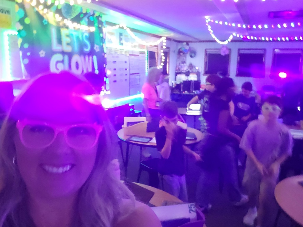 A woman with glasses smiles in a room with purple lighting. Behind her, several children and adults in the background.