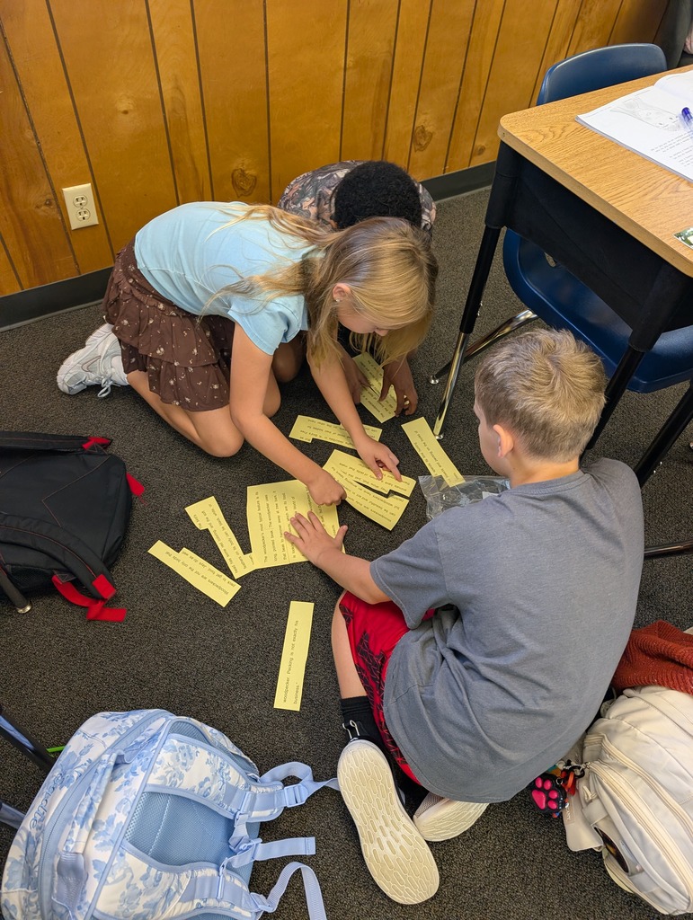 Children seated on the floor, sorting yellow cards, with backpacks and desks in a wooden-walled classroom.