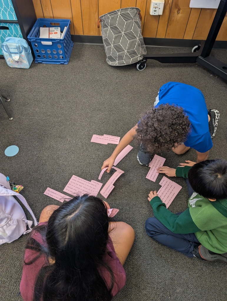 Three children work on a floor puzzle with pink cards, one with curly hair, another with a backpack.