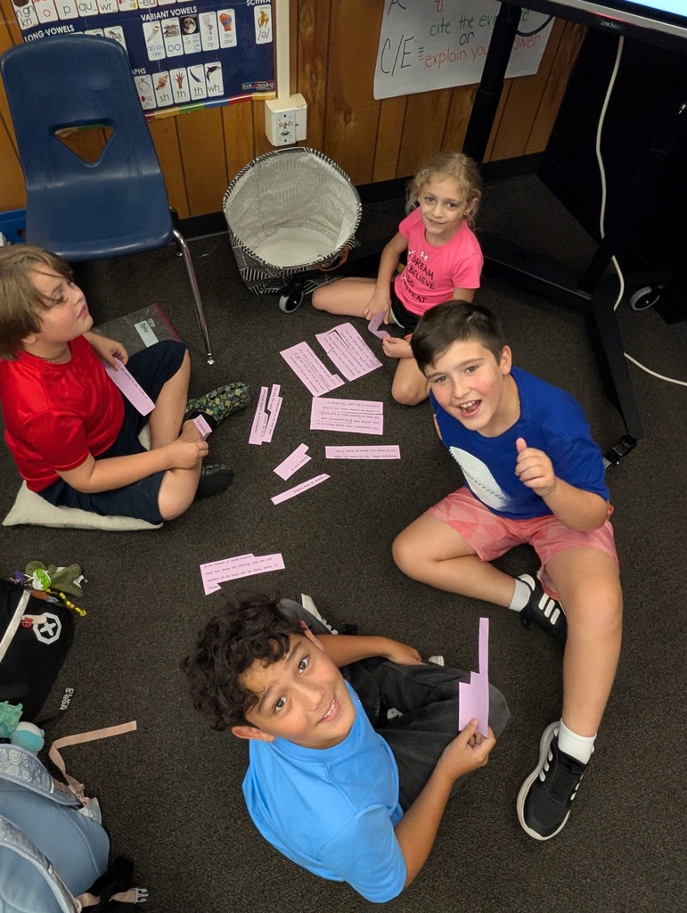 Four children sit on the floor, one holding a paper, others holding pink strips. A drum and chair are nearby.