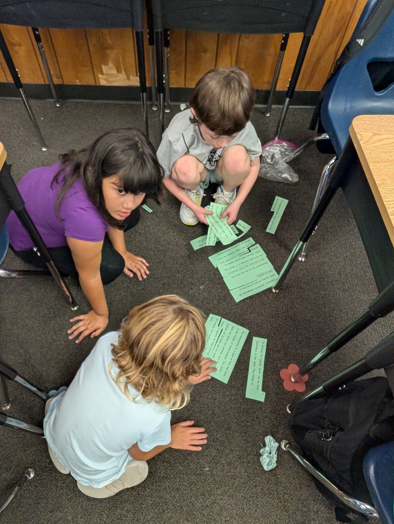 Children in a classroom crouch on the floor with papers and objects, focusing on a task.