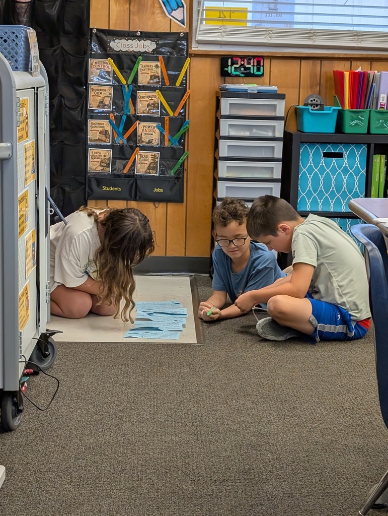 Three children sitting on floor, with one bending over to examine a worksheet. Shelves in background.