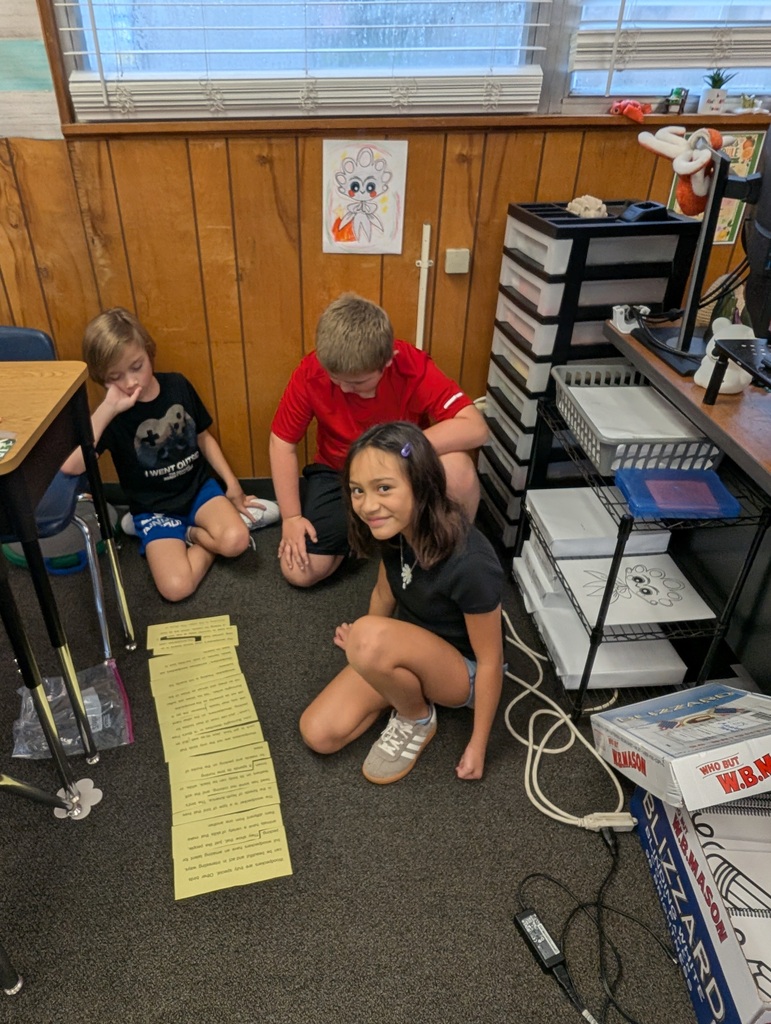 Three children work on floor in classroom. One leans on desk, one crouches with papers, and one stands behind them.