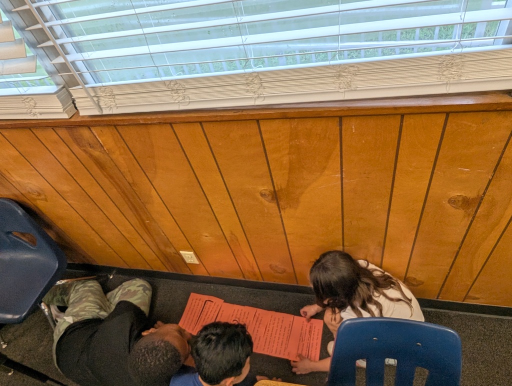Three children in a classroom, one sitting on the floor with a book, and two seated at a desk.