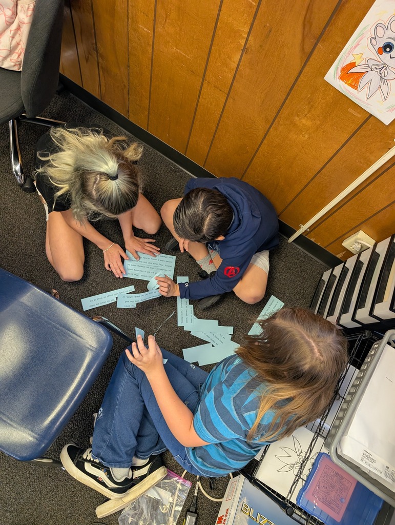 Three students are on the floor, working with paper. One student has blonde hair, and two have brown hair.