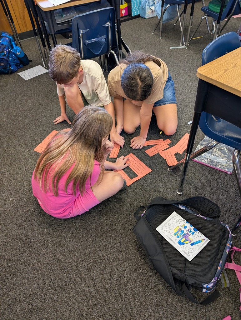 Three children seated on the floor work with colorful cards, with desks and chairs in a classroom.