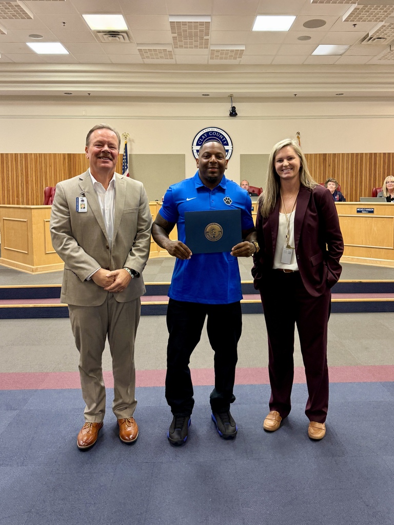 Three people stand on a blue carpet. The man in the center holds a certificate, and the two others wear business attire. Behind them is a wooden bench.