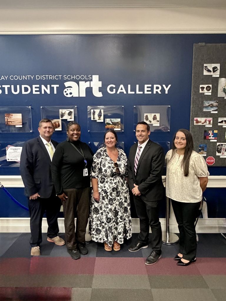 Five adults stand in front of a blue wall with "Student Art Gallery" in white letters. Some wear formal attire. Artworks and photographs adorn the wall behind them.
