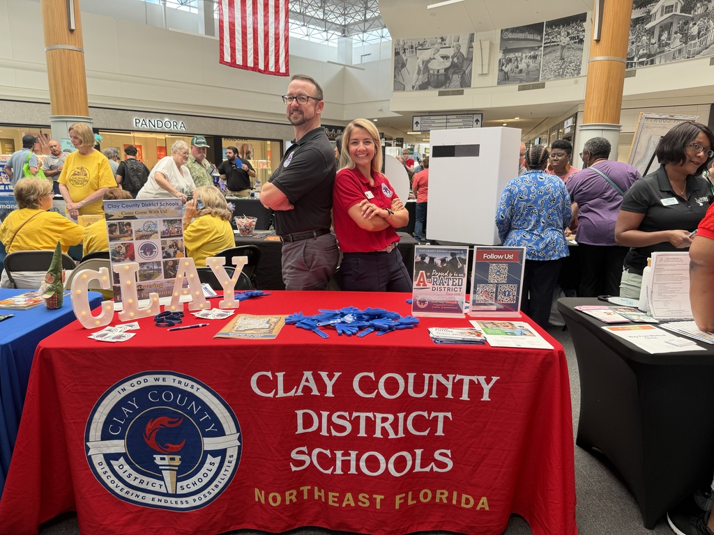 Two adults stand behind a table with a red cover. It reads Clay County District Schools Northeast Florida. People stand in the background.