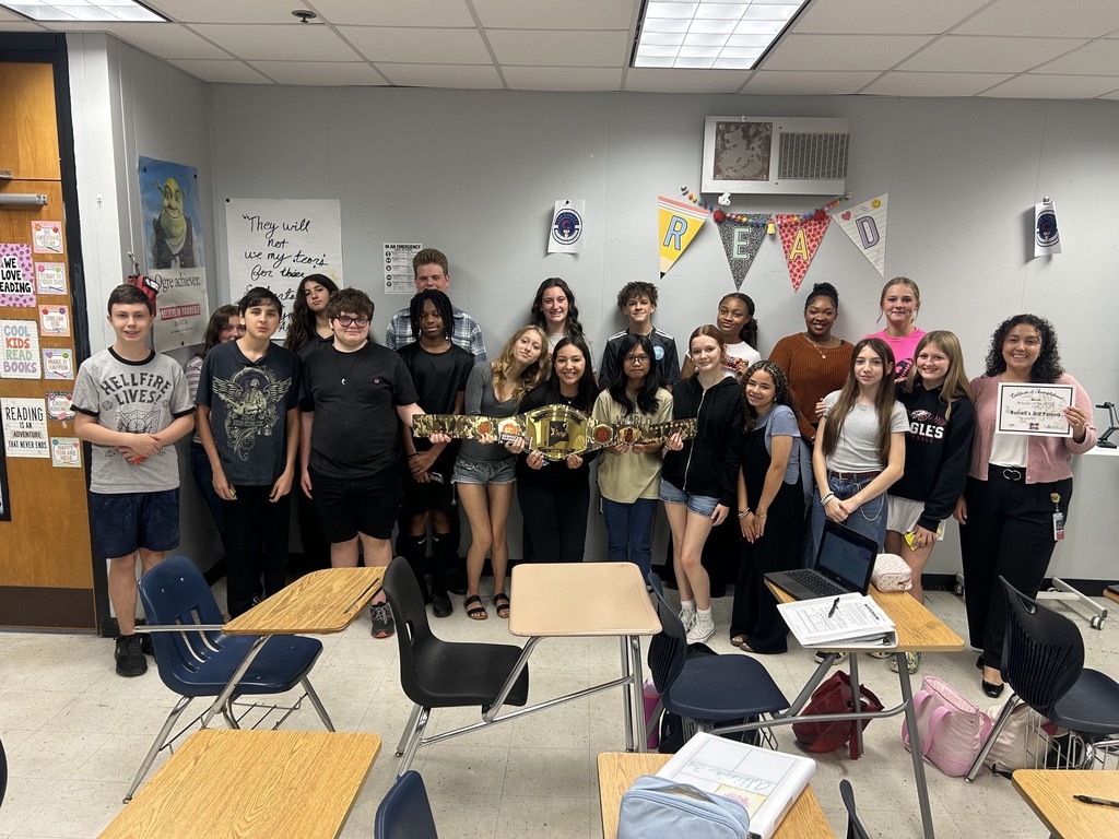 Group of students in a classroom with desks and chairs; some hold posters and one holds a trophy.