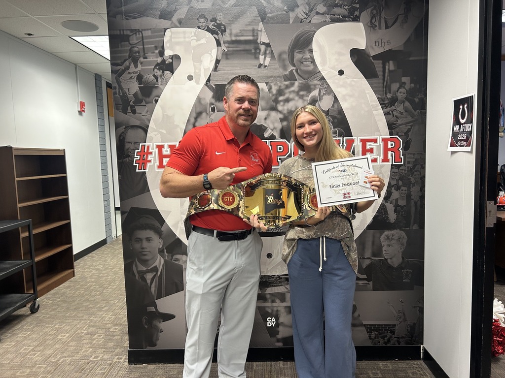 Man and woman stand, holding a championship belt and certificate, in an office with a wall mural.