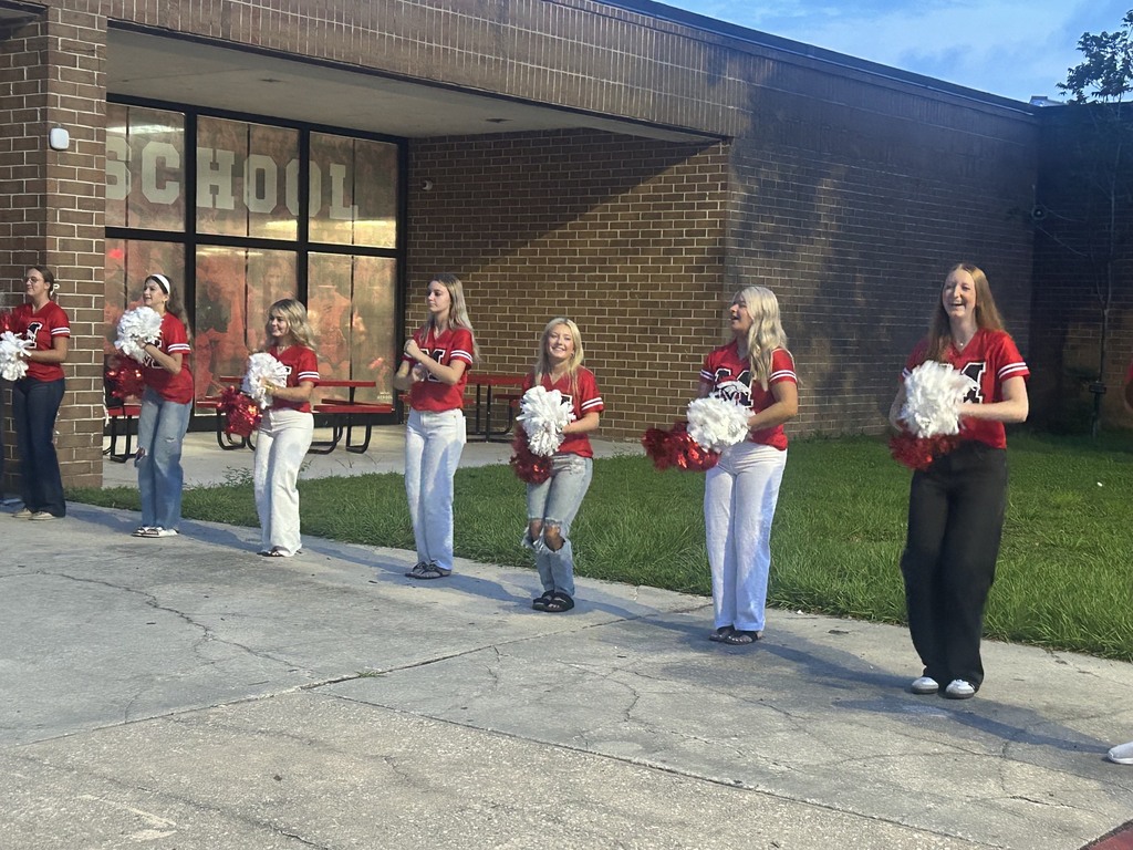 Seven cheerleaders in red shirts and white pants stand on a sidewalk, holding white pompoms, in front of a brick school building.