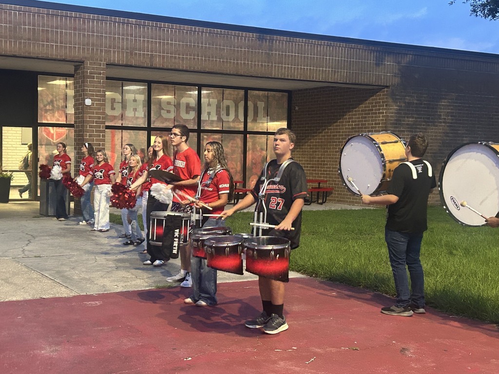 A group of students wearing red and white uniforms play drums in front of a brick school building.
