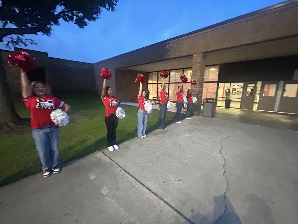 Cheerleaders in red uniforms stand on a sidewalk with pom-poms, near a building with glass windows.