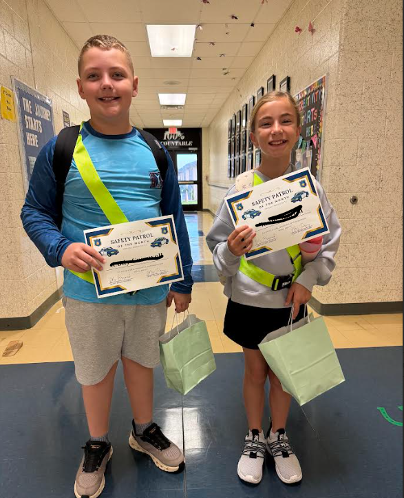 Two children stand in a hallway, holding certificates and bags, smiling brightly. The boy wears a blue shirt, and the girl wears a white sweatshirt.