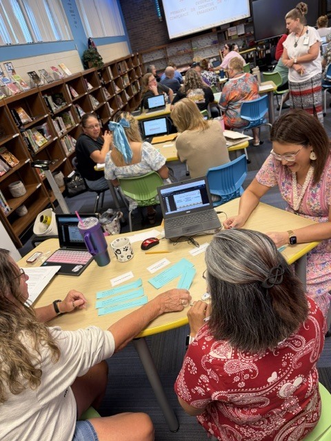 A group of adults at a table in a classroom setting, seated around a table with laptops and a projector screen.