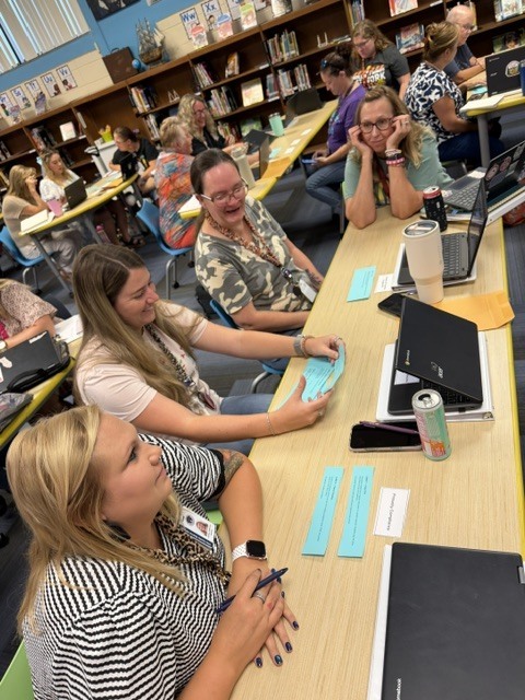 Several women sit at a table in a classroom, using laptops and papers. Some hold pens.