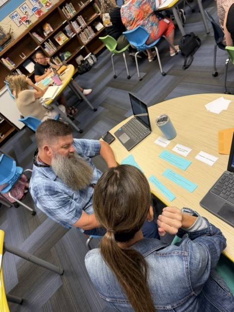 A person with a beard sits at a table with a woman, both working on laptops. Behind them, others sit at tables in a library.