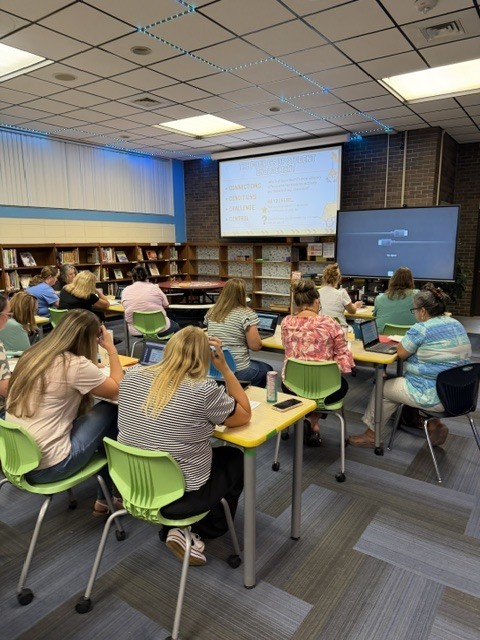 People sit around tables in a room with bookshelves and a projector screen on a wall.