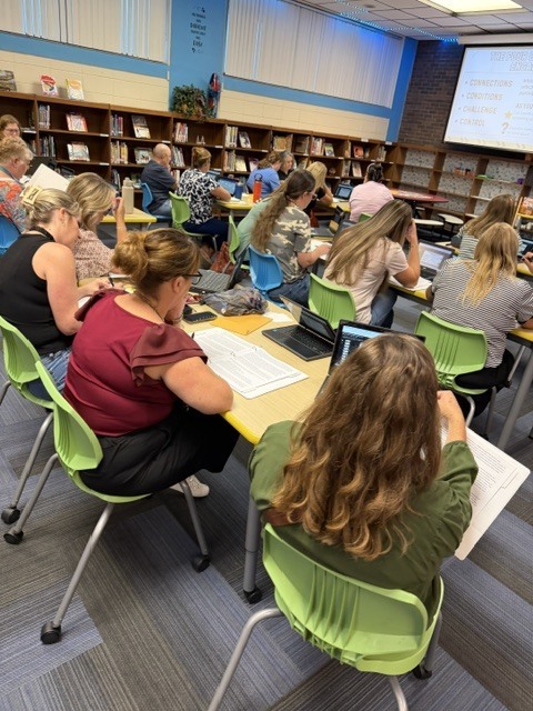 People in green chairs are sitting at tables in a library, using laptops, and looking at a projector screen.