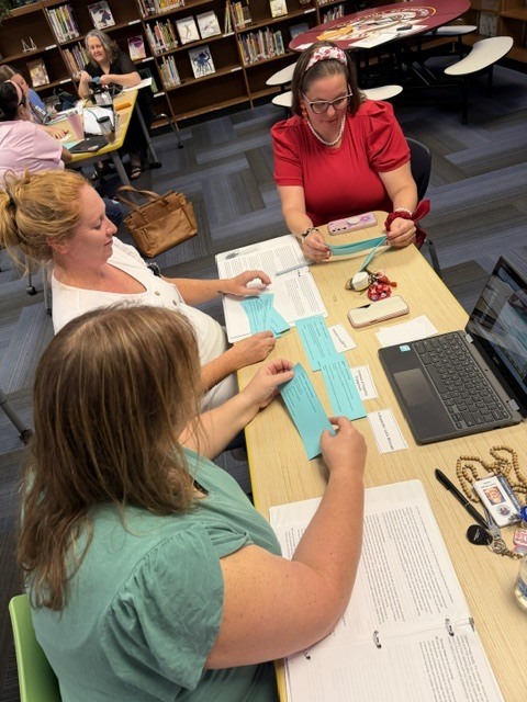 Three women in a library work at a table with a laptop and papers. They have bags and scissors.