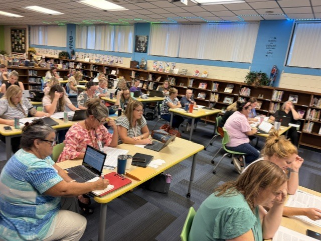 A room with rows of people seated at tables with laptops and books, a library in the background.