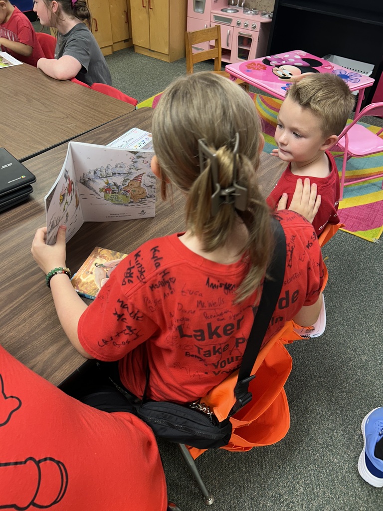 Girl reads a book to a boy sitting next to her at a table in a classroom.