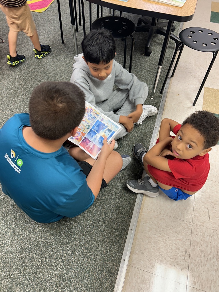 Three children are seated on the floor, reading a book. One boy wears a blue shirt, another in gray, and the third in red.